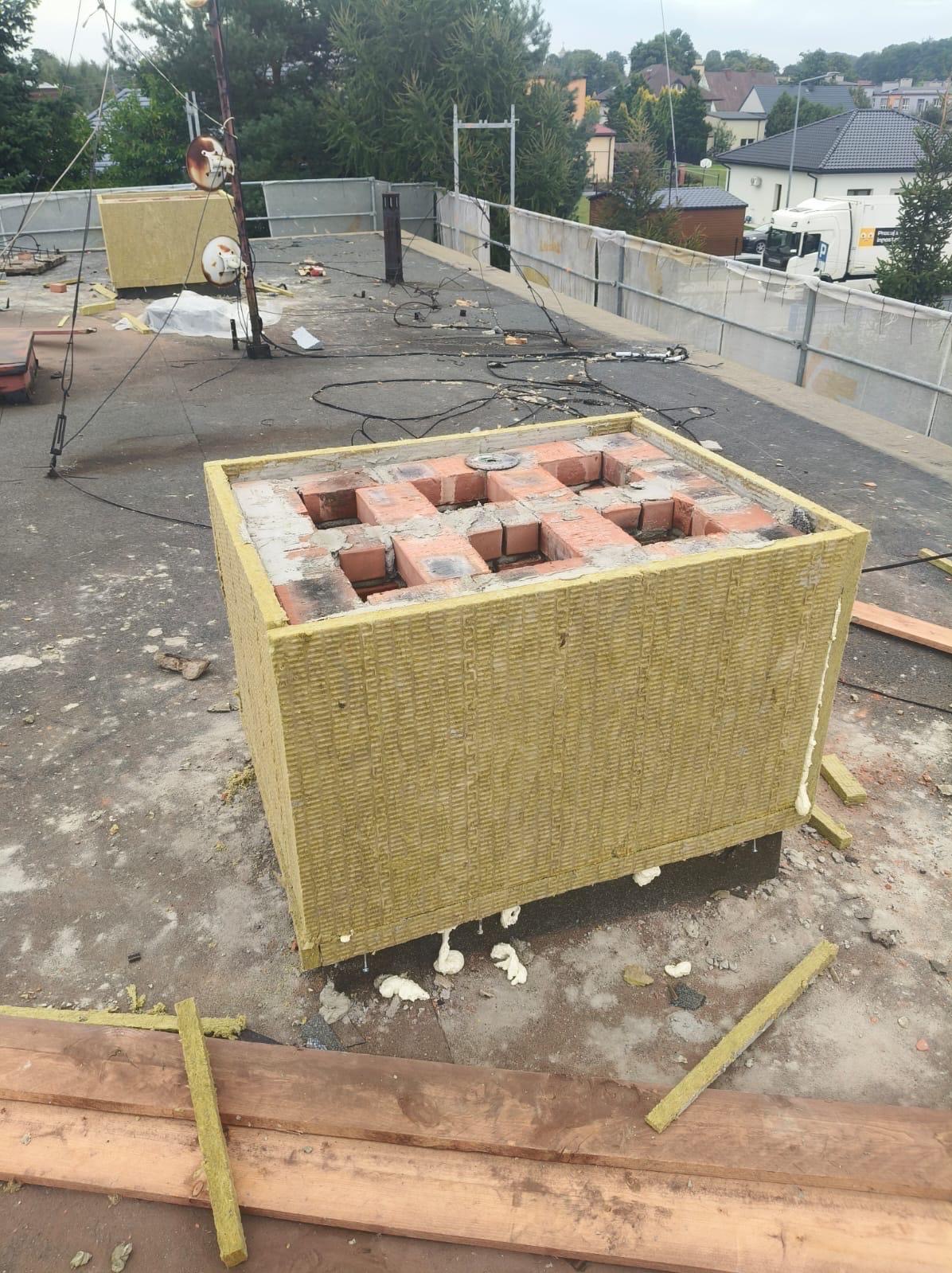 Ocieplony komin z cegły na dachu budynku, widoczne materiały izolacyjne i fragmenty konstrukcji. Prace na wysokości, w tle zabudowa mieszkalna.
