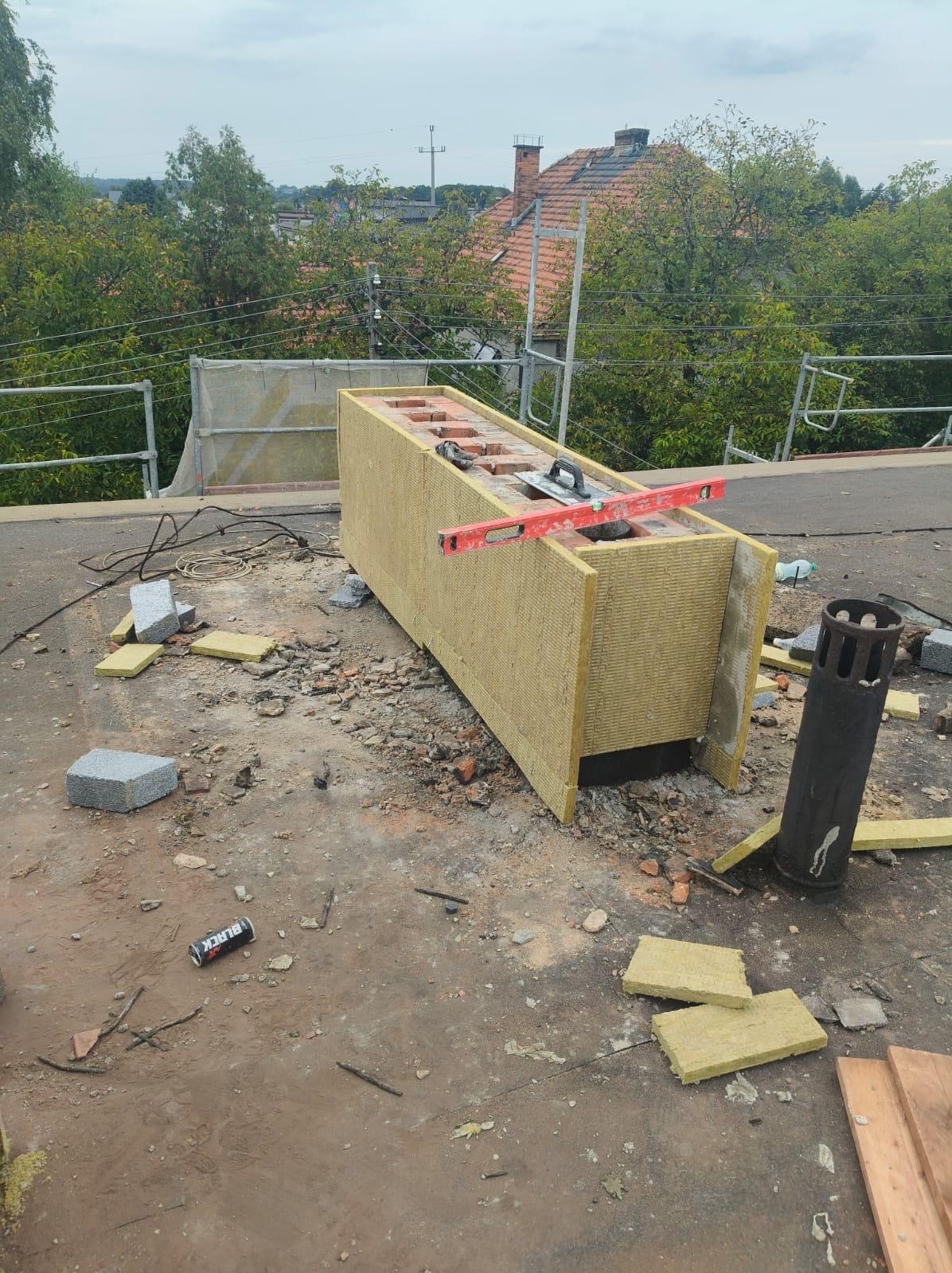 Ocieplony komin na dachu budynku w trakcie prac. Widoczne cegły, wełna mineralna, poziomica, gruz i puszka po napoju energetycznym. W tle zieleń i zabudowania.