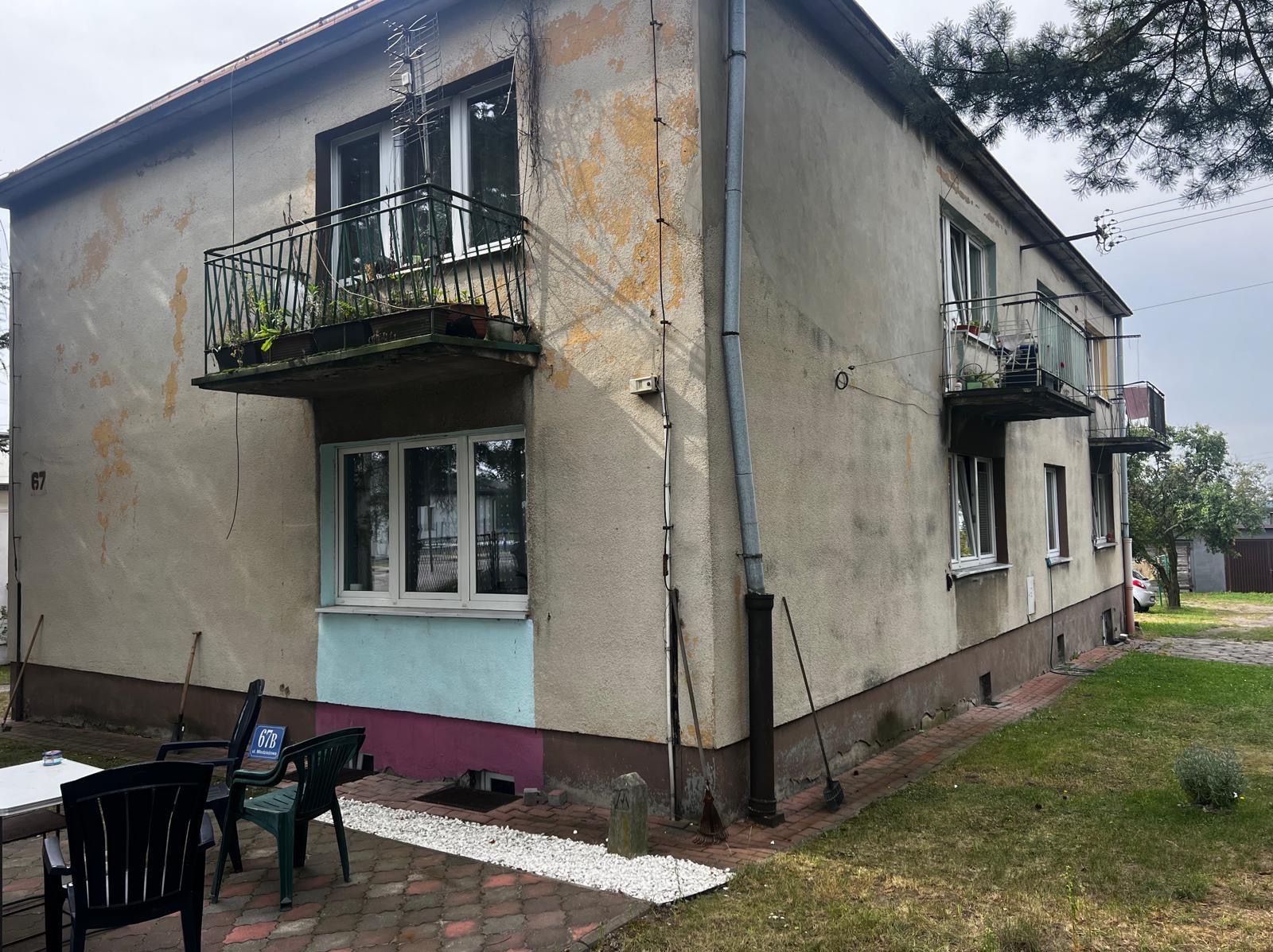 Dwupiętrowy budynek z balkonami, z widocznymi śladami wcześniejszych prac malarskich na elewacji. Fragment pomalowany na niebiesko i różowo. Widoczne krzesła ogrodowe.