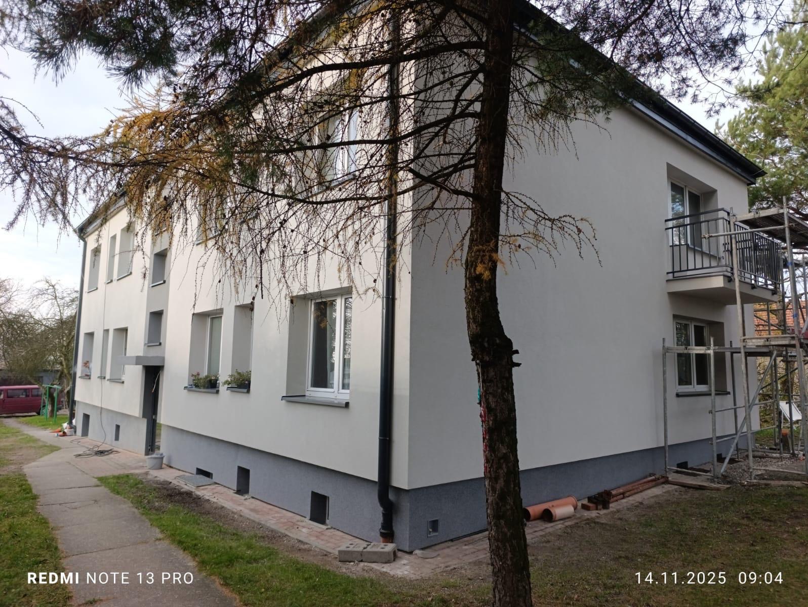 Odnowiona elewacja budynku wielorodzinnego z szarą podmurówką i białymi ścianami, widoczna z boku, z drzewem na pierwszym planie i rusztowaniem przy balkonie.