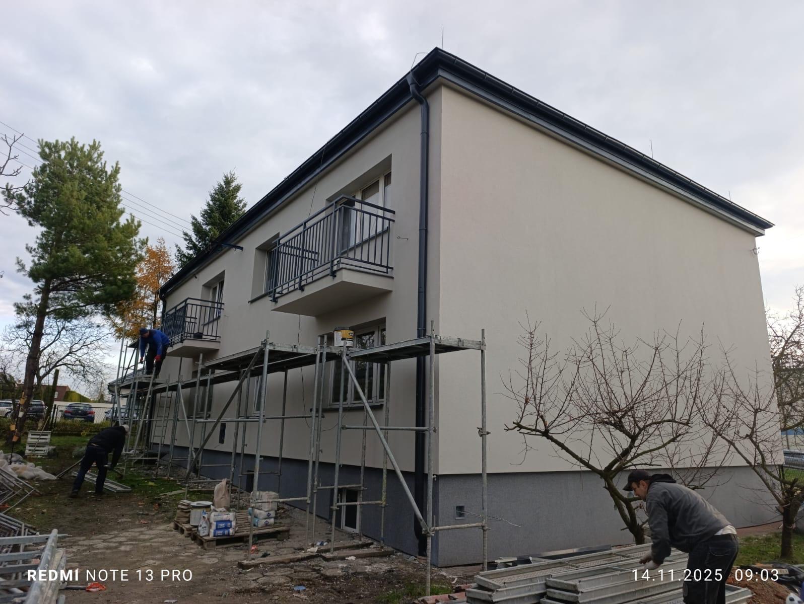 Dwupiętrowy budynek po renowacji elewacji, z balkonami i rusztowaniami. Pracownicy na rusztowaniach, widoczne materiały budowlane. Szara, pochmurna pogoda.