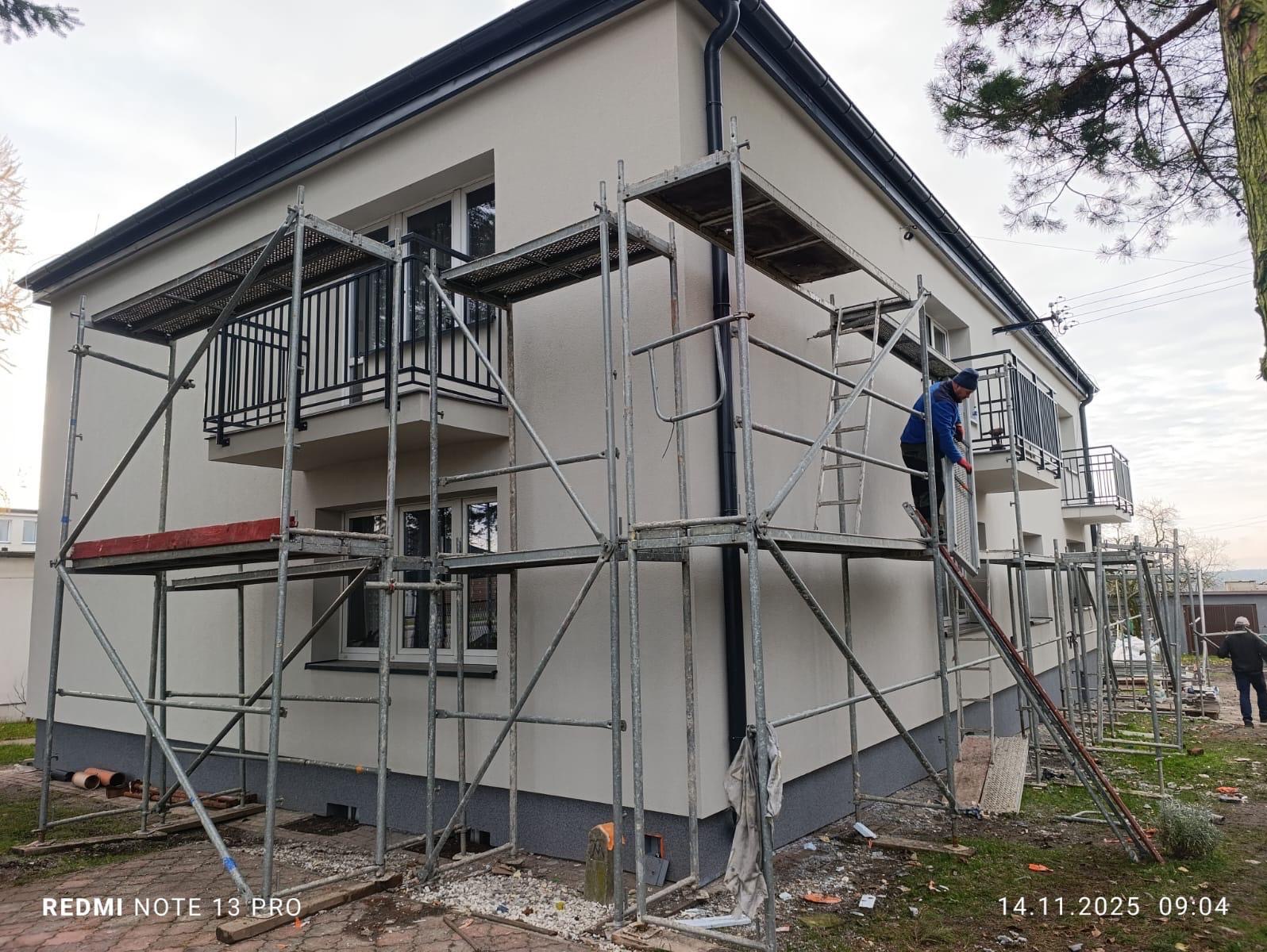 Dwupiętrowy budynek w trakcie prac elewacyjnych, otoczony rusztowaniami. Pracownik na drabinie, świeżo otynkowana ściana w jasnym odcieniu. Widoczne balkony z czarną balustradą.