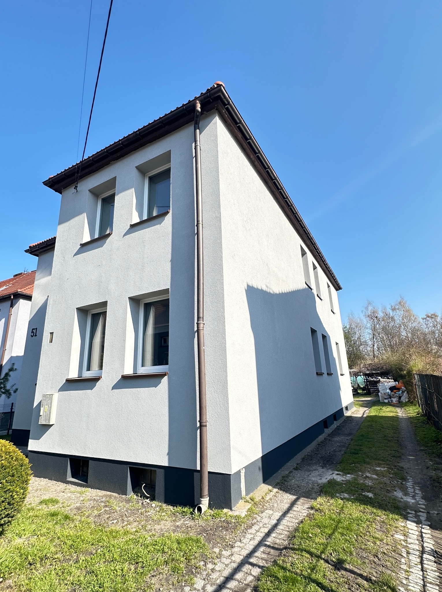 Odnowiona elewacja dwukondygnacyjnego domu z numerem 51, pomalowana na szaro, z ciemniejszym cokołem. Widoczne okna z białymi ramami i brązowa rynna.