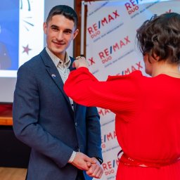 Agent nieruchomości Maciej Tomalak - Agent nieruchomości RE/MAX odbiera wyróżnienie. Kobieta w czerwonej sukience przypina odznakę do klapy marynarki uśmiechniętego mężczyzny na tle logotypu firmy.