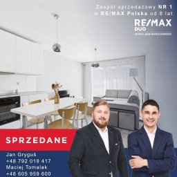 Agent nieruchomości Maciej Tomalak - Jasne wnętrze mieszkania z jadalnią i salonem, oznaczone jako sprzedane. W tle widoczni dwaj agenci nieruchomości RE/MAX Duo.