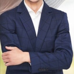 Agent nieruchomości Maciej Tomalak - Uśmiechnięty agent nieruchomości w granatowej marynarce w kratę, białej koszuli i bordowych spodniach, z założonymi rękami, na rozmytym tle.