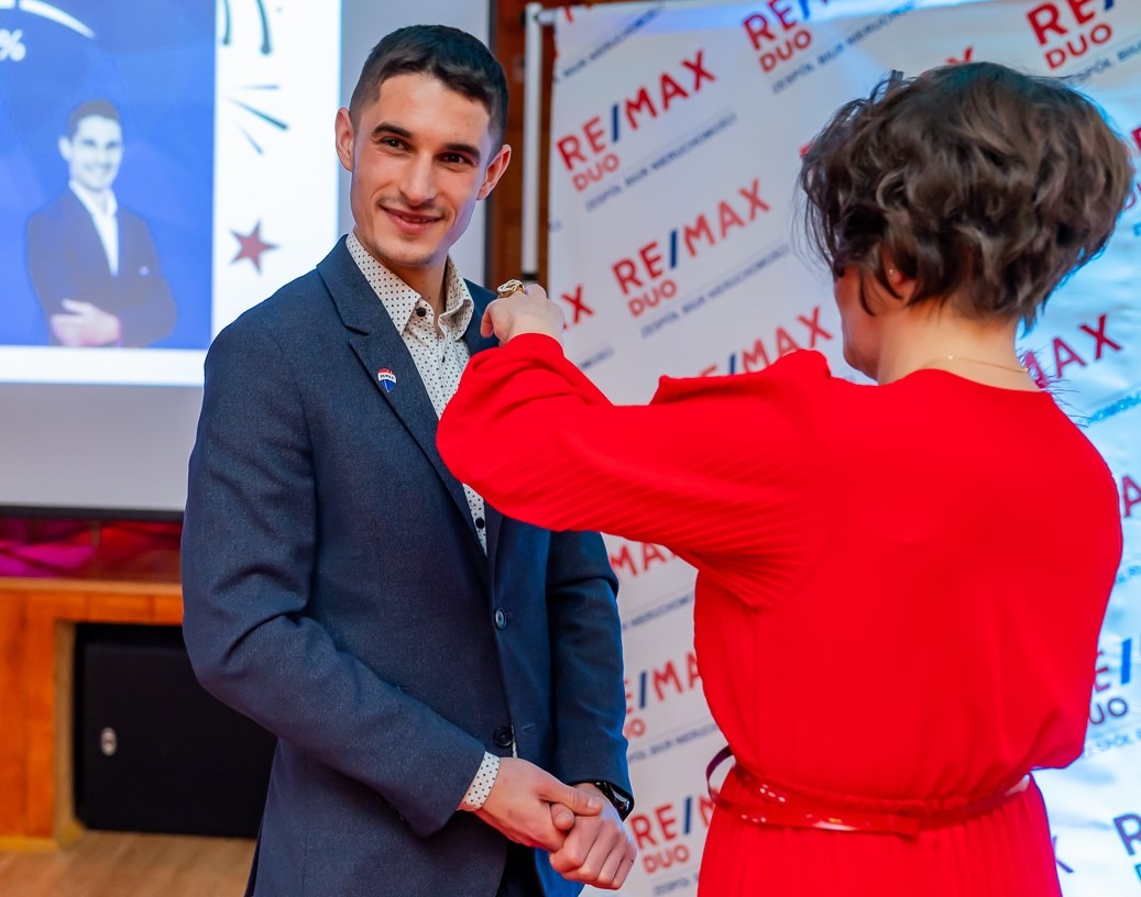 Agent nieruchomości RE/MAX odbiera wyróżnienie. Kobieta w czerwonej sukience przypina odznakę do klapy marynarki uśmiechniętego mężczyzny na tle logotypu firmy.