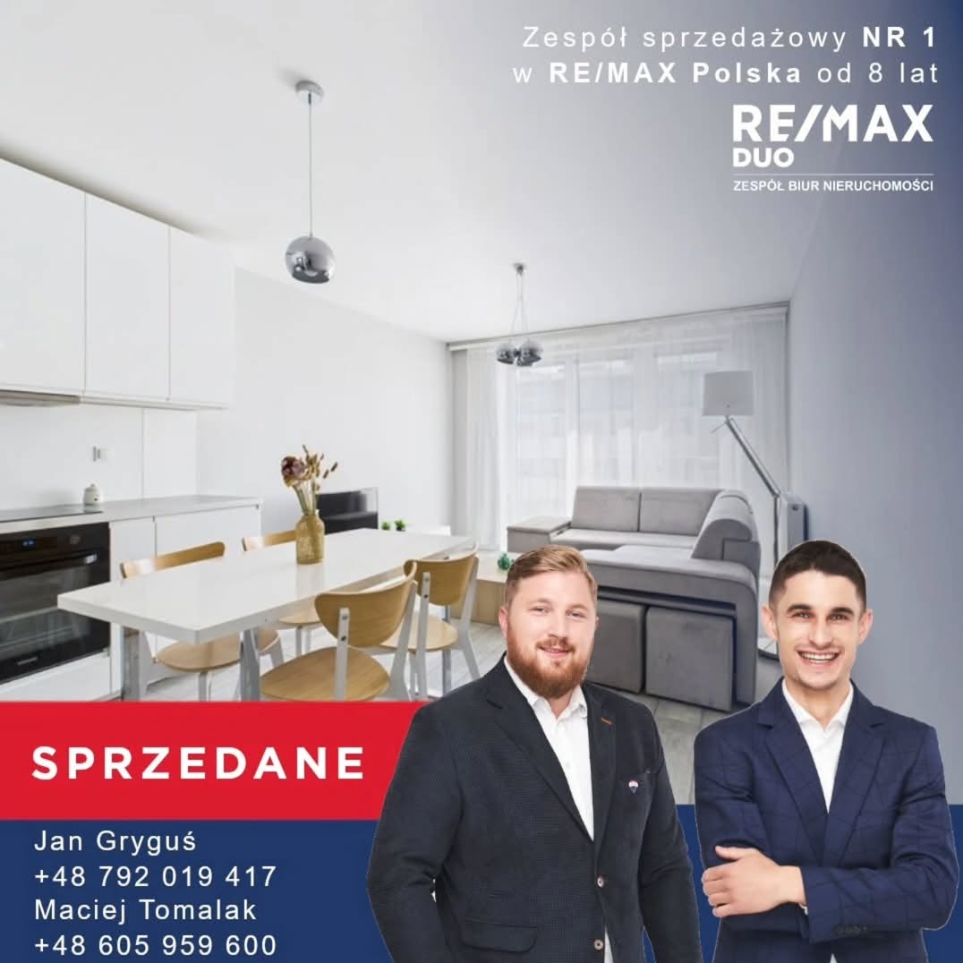 Jasne wnętrze mieszkania z jadalnią i salonem, oznaczone jako sprzedane. W tle widoczni dwaj agenci nieruchomości RE/MAX Duo.