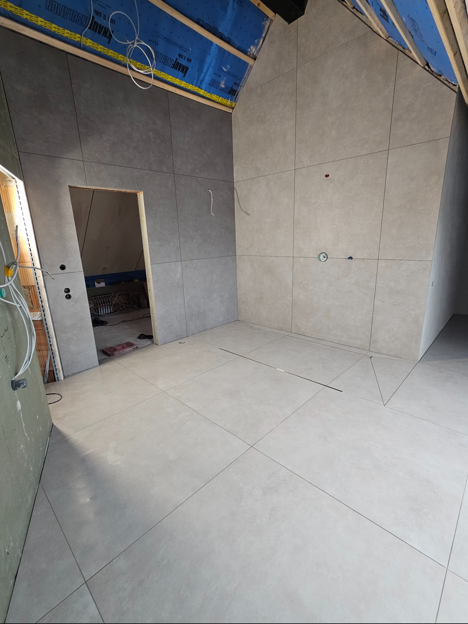 Wykończona łazienka z szarymi płytkami wielkoformatowymi na ścianach i podłodze. Widoczne pionowe i poziome fugi. W tle surowy strop i instalacje.