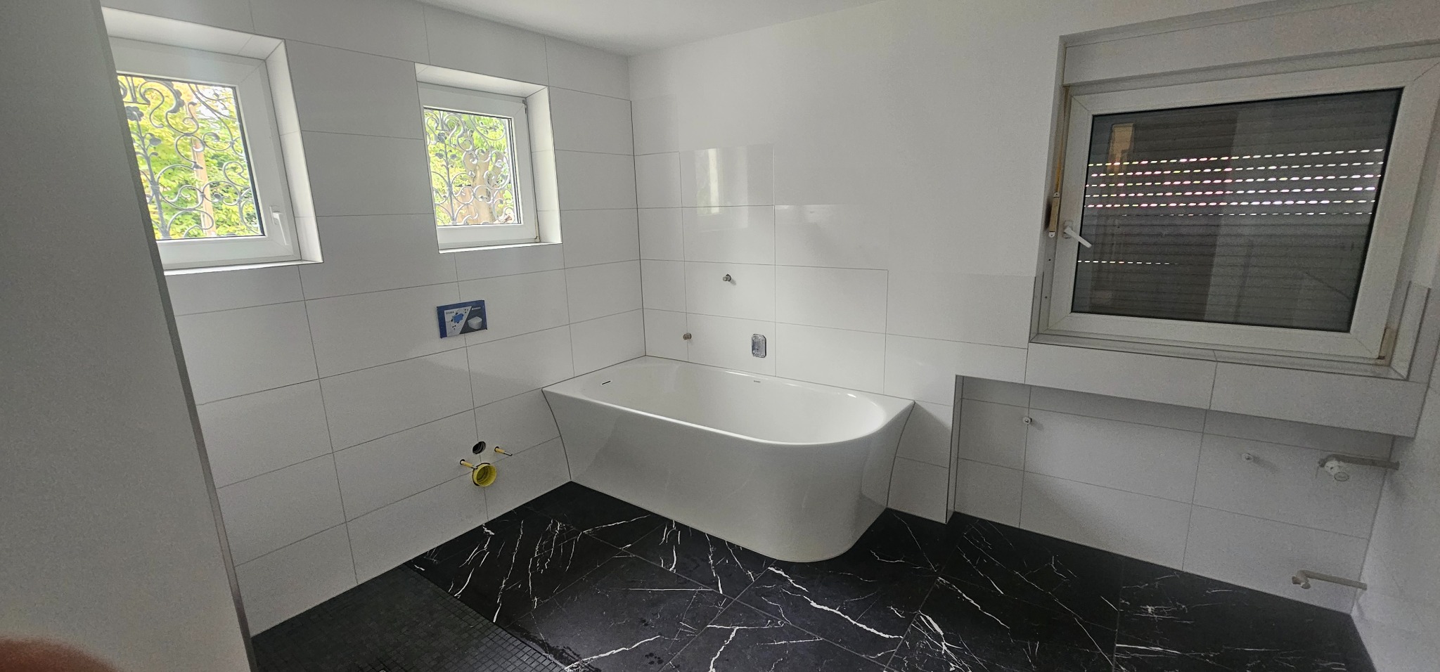 Nowoczesna łazienka z białą wanną wolnostojącą i czarną marmurową podłogą. Białe płytki na ścianach, okna z ozdobnymi kratami i roletami. Widoczne przyłącza hydrauliczne.