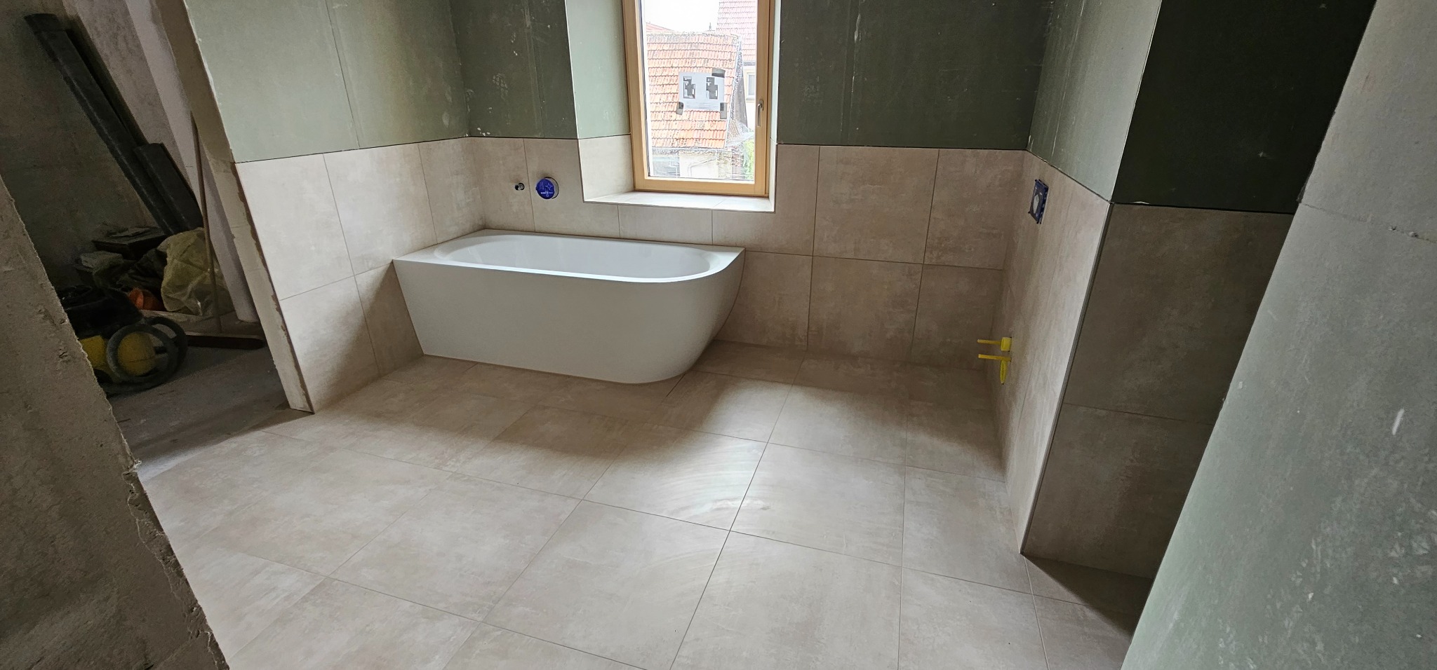 Jasna łazienka w trakcie wykończenia: beżowe płytki na podłodze i ścianach do połowy wysokości, wolnostojąca wanna przy oknie, ściany częściowo wykończone płytą gipsową.