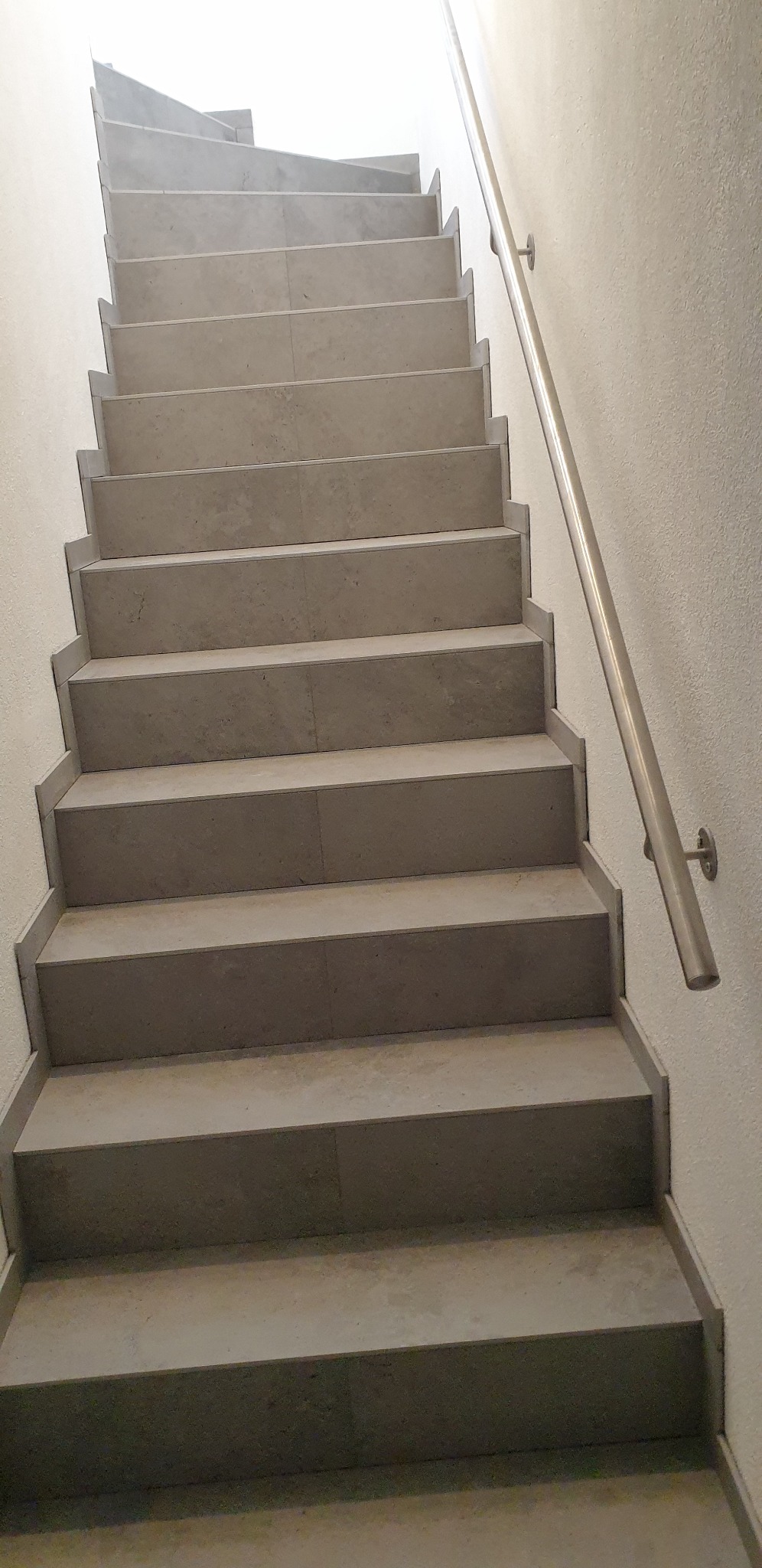 Schody wyłożone płytkami w odcieniach szarości, z metalową poręczą po prawej stronie, oświetlone od góry, widziane z dołu, z perspektywą w górę.