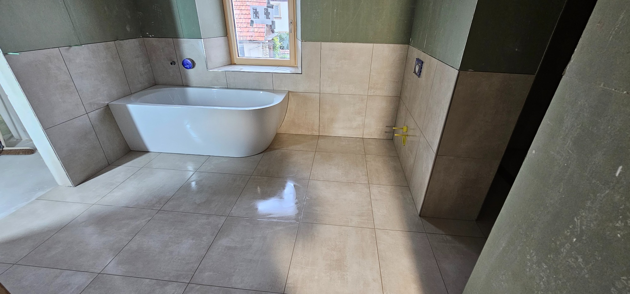 Jasna łazienka w trakcie wykańczania: beżowe płytki na podłodze i ścianach, biała wanna, okno z widokiem na budynek. Widoczne elementy instalacji.