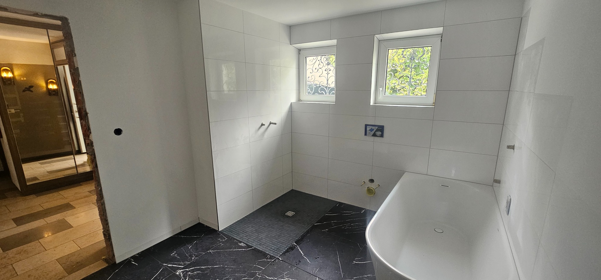 Biała łazienka z wanną i kabiną prysznicową, wykończona płytkami. Widoczne dwa okna z ozdobnymi kratami i czarna podłoga imitująca marmur.