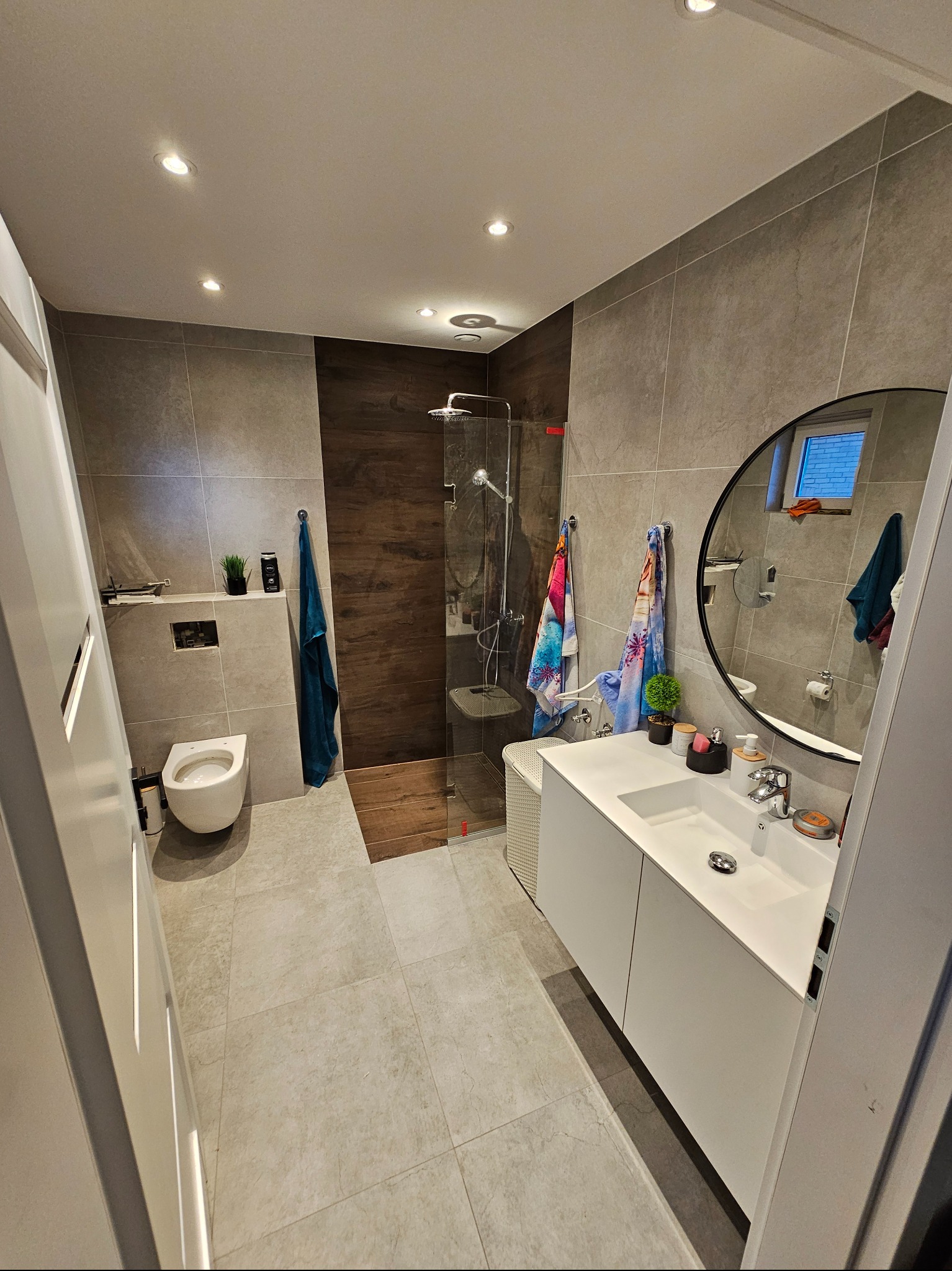 Nowoczesna łazienka z kabiną prysznicową typu walk-in, umywalką w zabudowie i okrągłym lustrem. Szare płytki i drewnopodobne akcenty. Oświetlenie punktowe.