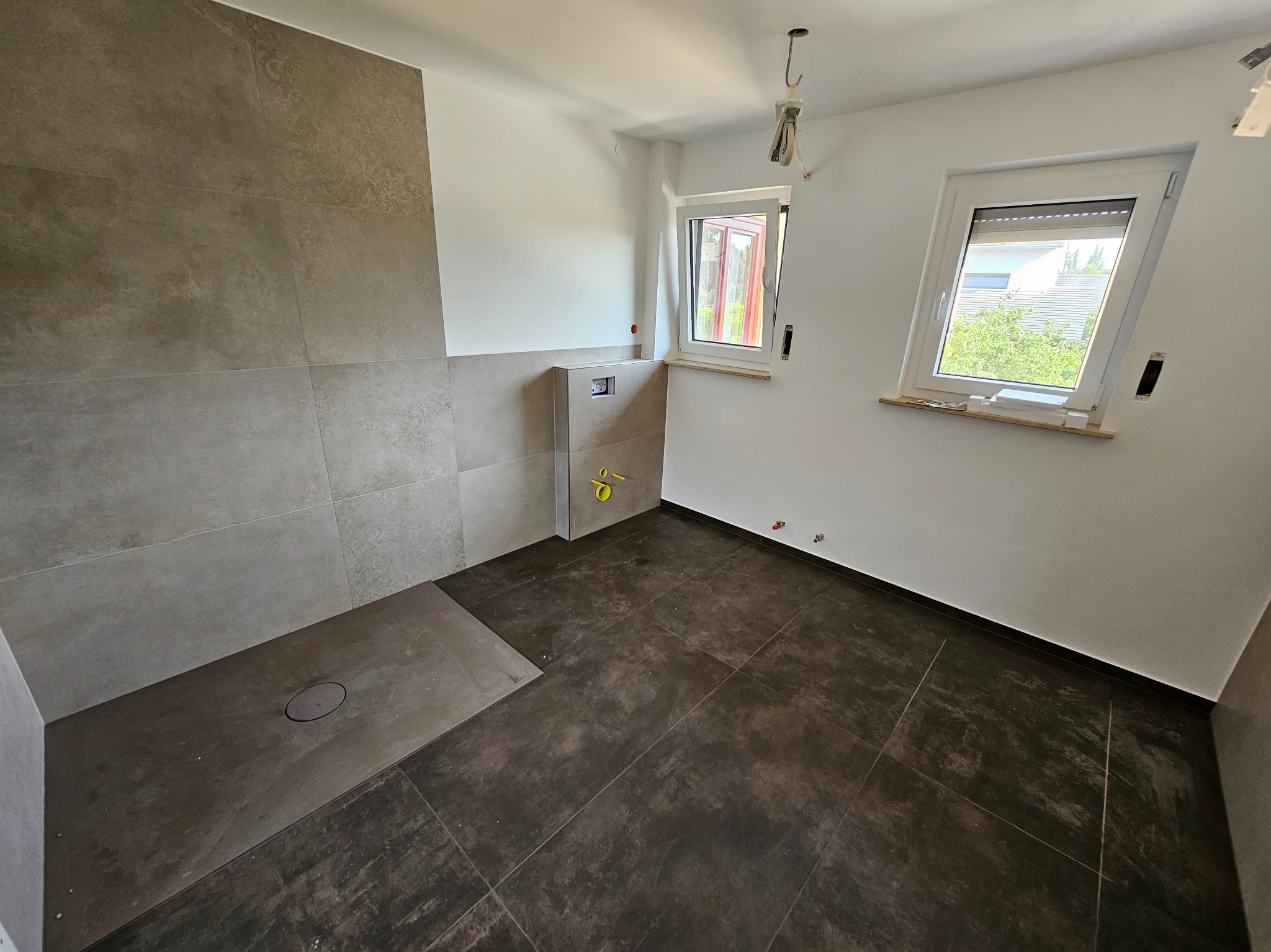 Wykończona łazienka z brązowymi płytkami podłogowymi i szarą strefą prysznicową. Widoczne okna i instalacje. Minimalistyczny design z elementami w trakcie montażu.