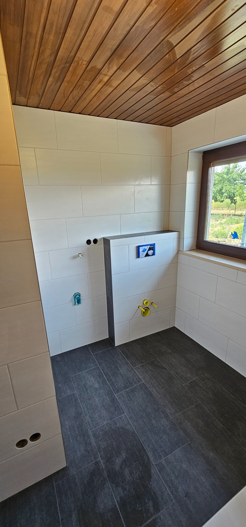 Wykończona łazienka z drewnianym sufitem, białymi płytkami ściennymi i ciemnymi płytkami podłogowymi. Widoczne przyłącza wodne i okno z widokiem na zieleń.