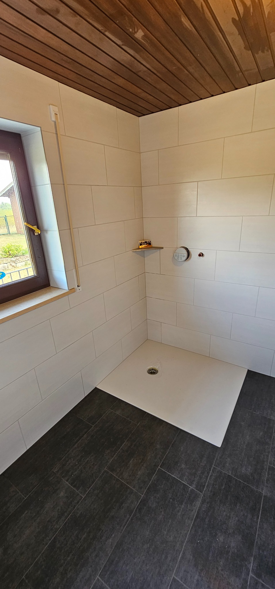 Wykończona kabina prysznicowa z białymi płytkami ściennymi i ciemną podłogą z płytek imitujących kamień. Widoczny drewniany sufit i okno z brązową ramą.