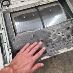 MAGIK AGD - Wnętrze pralki z nagromadzeniem szarego pyłu i włókien, dłoń wskazuje na zanieczyszczenia. Widoczne elementy konstrukcyjne i przewody.