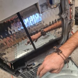 MAGIK AGD - Wnętrze pralki automatycznej z nagromadzonymi kłaczkami, widoczna dłoń usuwająca zanieczyszczenia. Perspektywa z góry, oświetlenie naturalne.