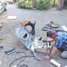 MAGIK AGD - Mężczyzna w koszuli w kratę naprawia rozłożoną pralkę na brukowanym podwórku. Widoczne części urządzenia, narzędzia i fragment samochodu w tle.