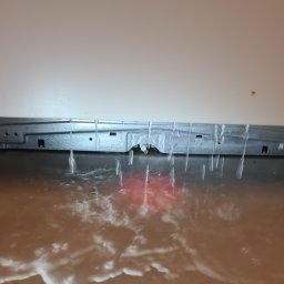 MAGIK AGD - Wyciekająca woda spod białej zmywarki, zalewająca podłogę. Widoczne metalowe elementy konstrukcyjne urządzenia. Awaria zmywarki.