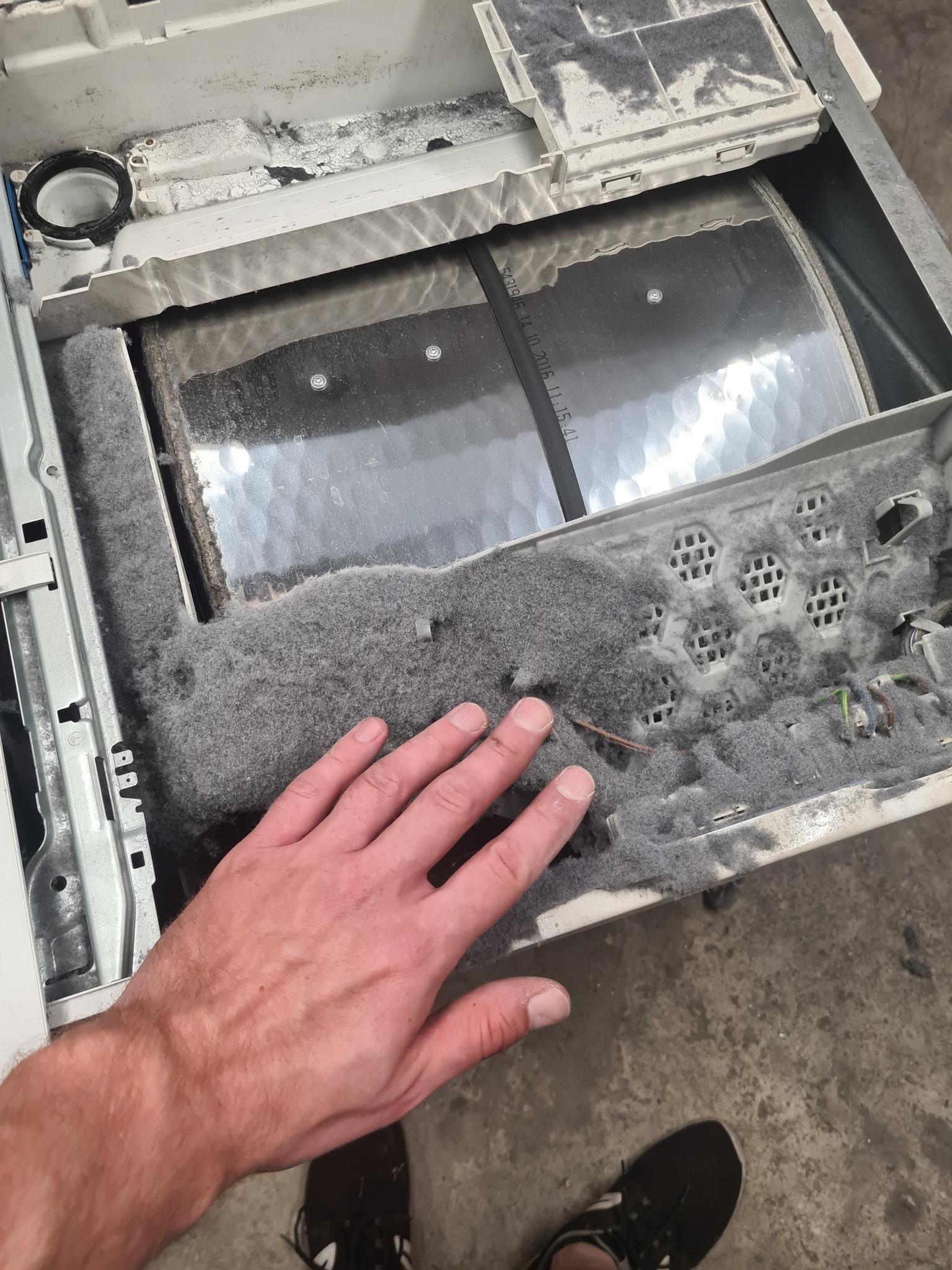 Wnętrze pralki z nagromadzeniem szarego pyłu i włókien, dłoń wskazuje na zanieczyszczenia. Widoczne elementy konstrukcyjne i przewody.