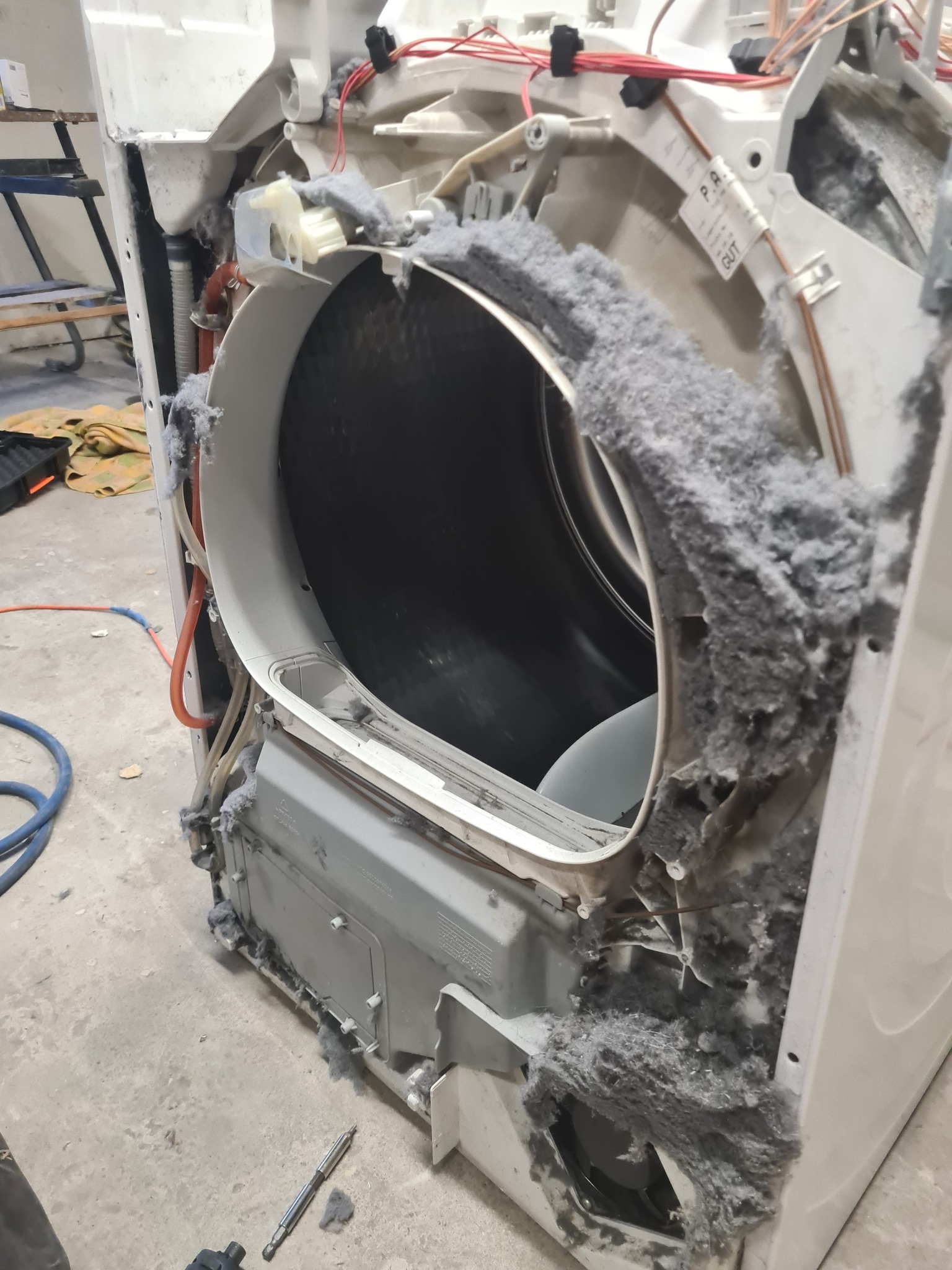 Otwarta pralka z widocznymi elementami wewnętrznymi i nagromadzeniem kurzu/włókien, sugerująca potrzebę czyszczenia lub naprawy. Widoczne narzędzia obok.