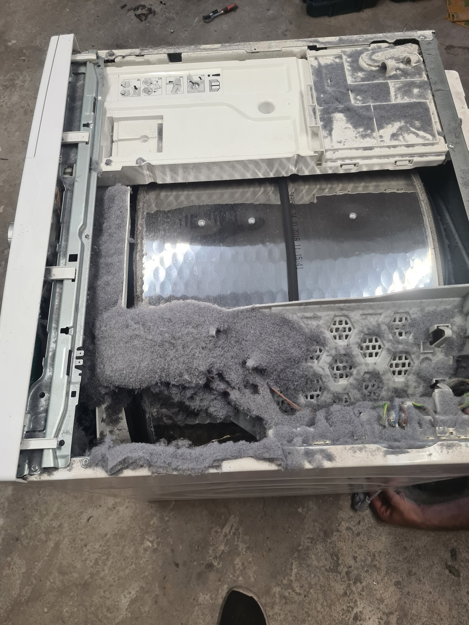 Otwarta pralka z nagromadzeniem szarego pyłu i włókien wewnątrz bębna, ukazująca stopień zużycia i potrzebę czyszczenia lub naprawy.