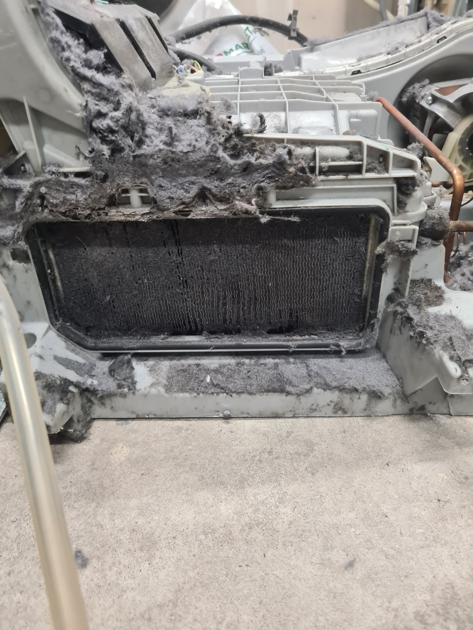 Zablokowany filtr suszarki bębnowej, oblepiony grubą warstwą szarego kurzu i włókien. Widoczne elementy konstrukcyjne urządzenia i fragment podłogi.
