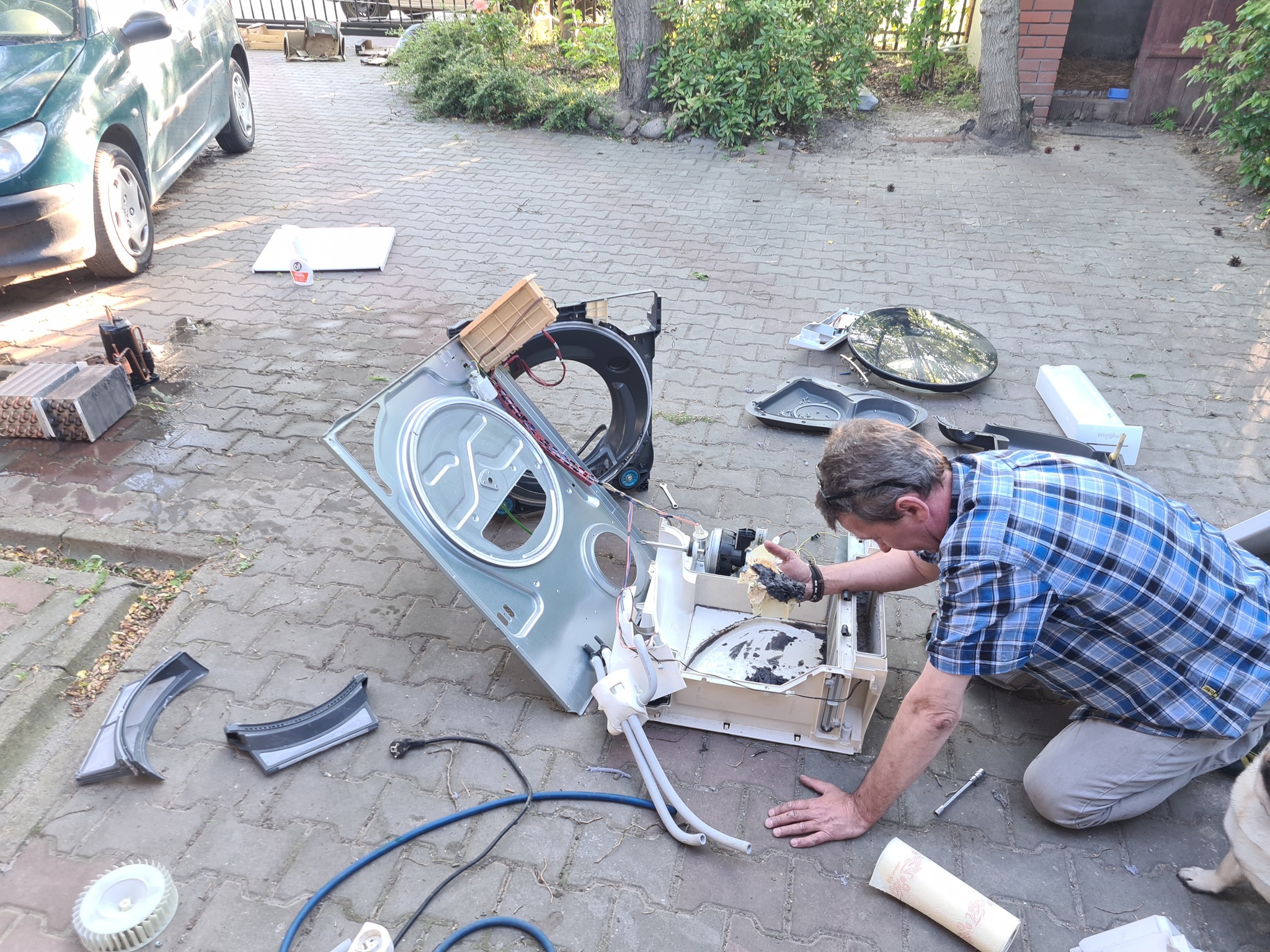 Mężczyzna w koszuli w kratę naprawia rozłożoną pralkę na brukowanym podwórku. Widoczne części urządzenia, narzędzia i fragment samochodu w tle.