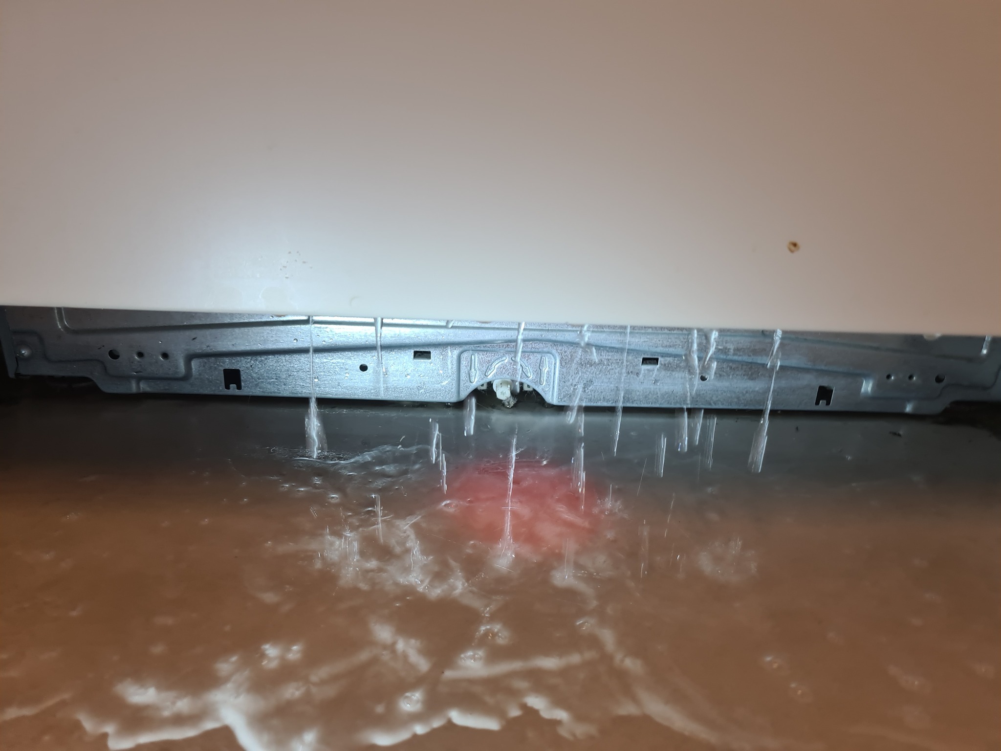 Wyciekająca woda spod białej zmywarki, zalewająca podłogę. Widoczne metalowe elementy konstrukcyjne urządzenia. Awaria zmywarki.