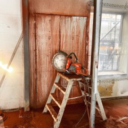 Pol - Cut Technika Diamentowa Jacek Suchodolski - Pomarańczowa piła do betonu Husqvarna na drabinie, ślady cięcia na ścianie i podłodze. Przygotowanie do wyburzania ściany, widoczne zabezpieczenia okna folią.