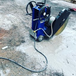 Pol - Cut Technika Diamentowa Jacek Suchodolski - Niebieska piła do cięcia betonu na mokro, z tarczą diamentową, stoi na placu budowy w kałuży. Widoczne przewody i elementy konstrukcyjne w tle.