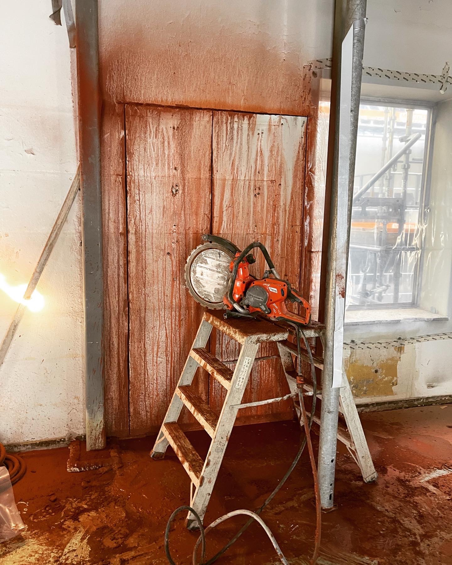 Pomarańczowa piła do betonu Husqvarna na drabinie, ślady cięcia na ścianie i podłodze. Przygotowanie do wyburzania ściany, widoczne zabezpieczenia okna folią.