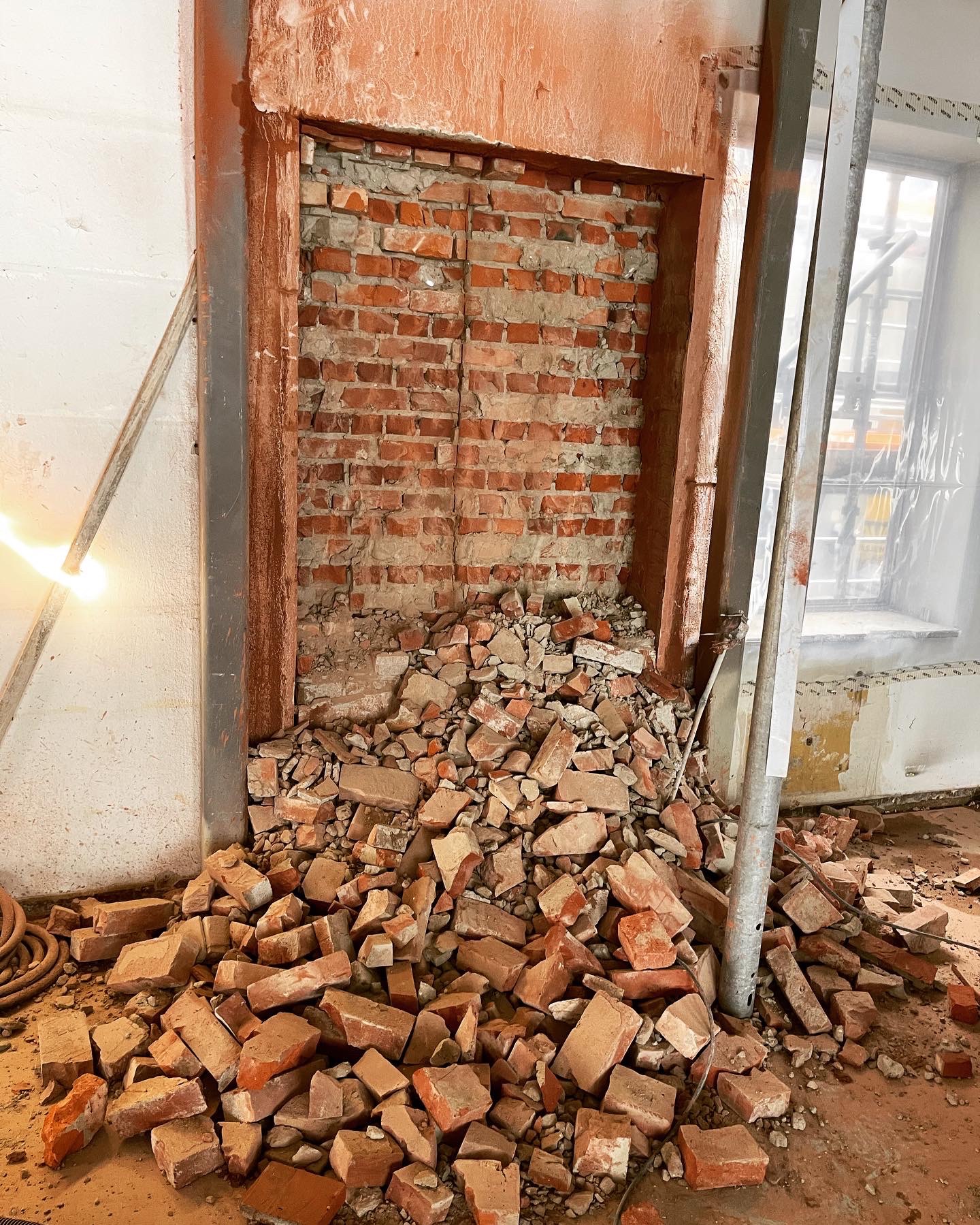 Ceglana ściana z otworem po usuniętej części, gruz na podłodze, metalowe wzmocnienia. Widoczne ślady po pracach demontażowych.