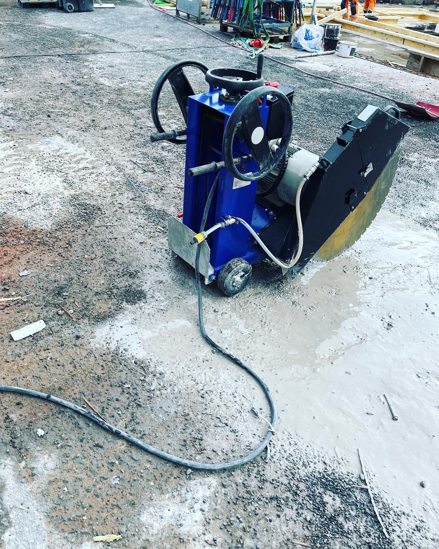 Niebieska piła do cięcia betonu na mokro, z tarczą diamentową, stoi na placu budowy w kałuży. Widoczne przewody i elementy konstrukcyjne w tle.