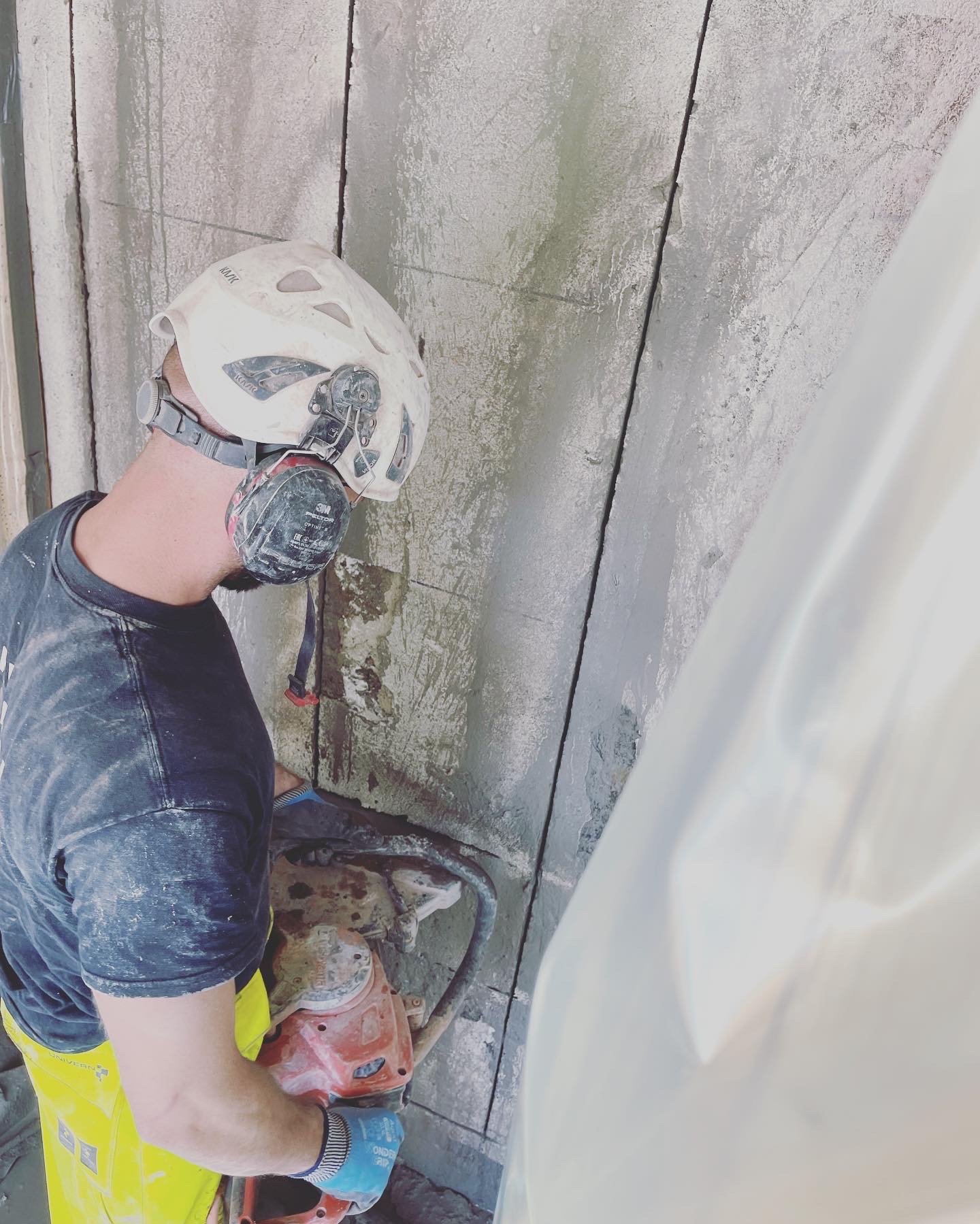Pracownik w kasku i ochronnikach słuchu, z maską na twarzy, używa piły do cięcia betonu na ścianie. Widoczne ślady cięcia i pył. Częściowo zasłonięte folią ochronną.