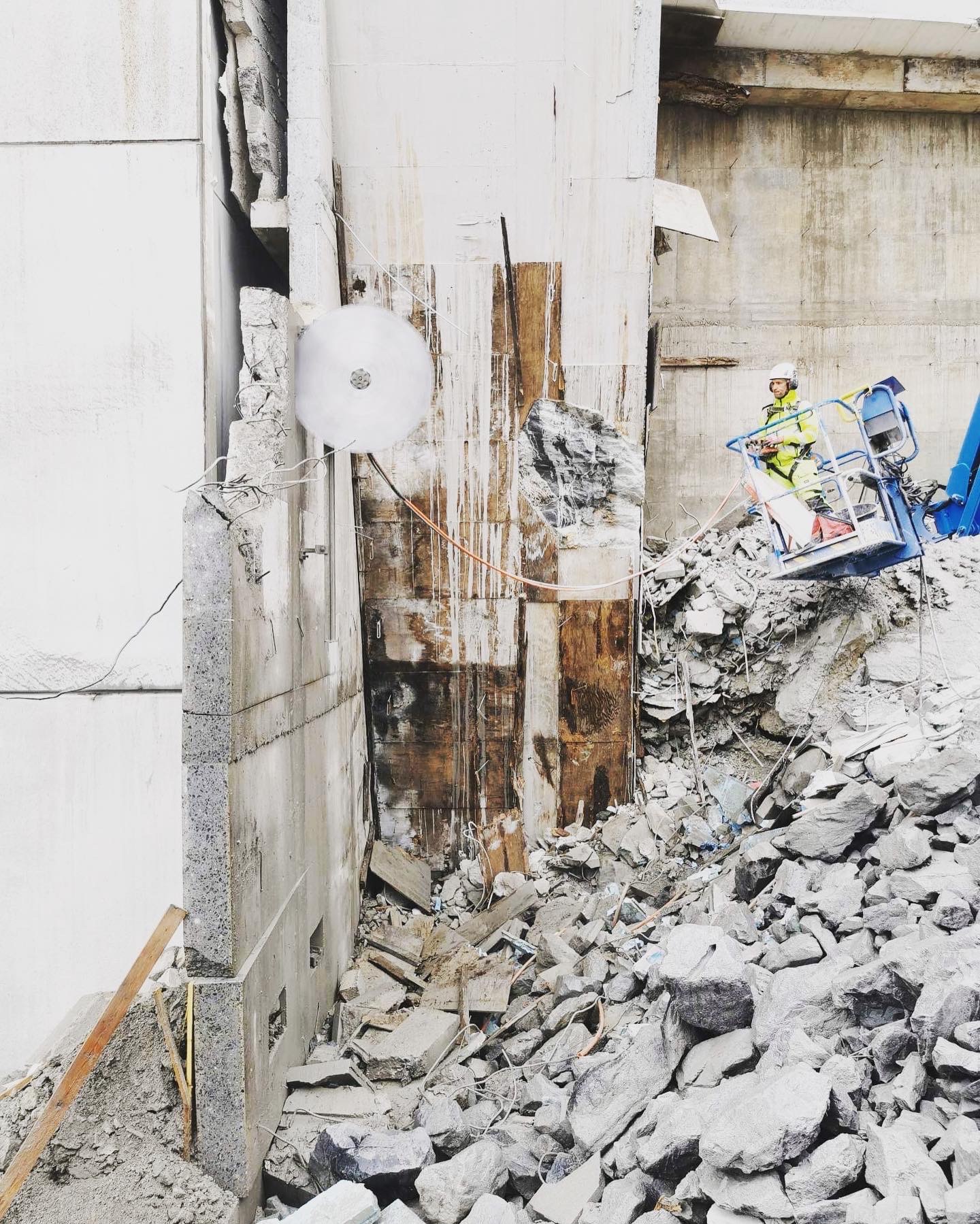 Pracownik w podnośniku koszowym tnie betonową ścianę tarczą diamentową. Gruz i fragmenty ściany na ziemi. Widoczne zbrojenia i elementy konstrukcyjne.