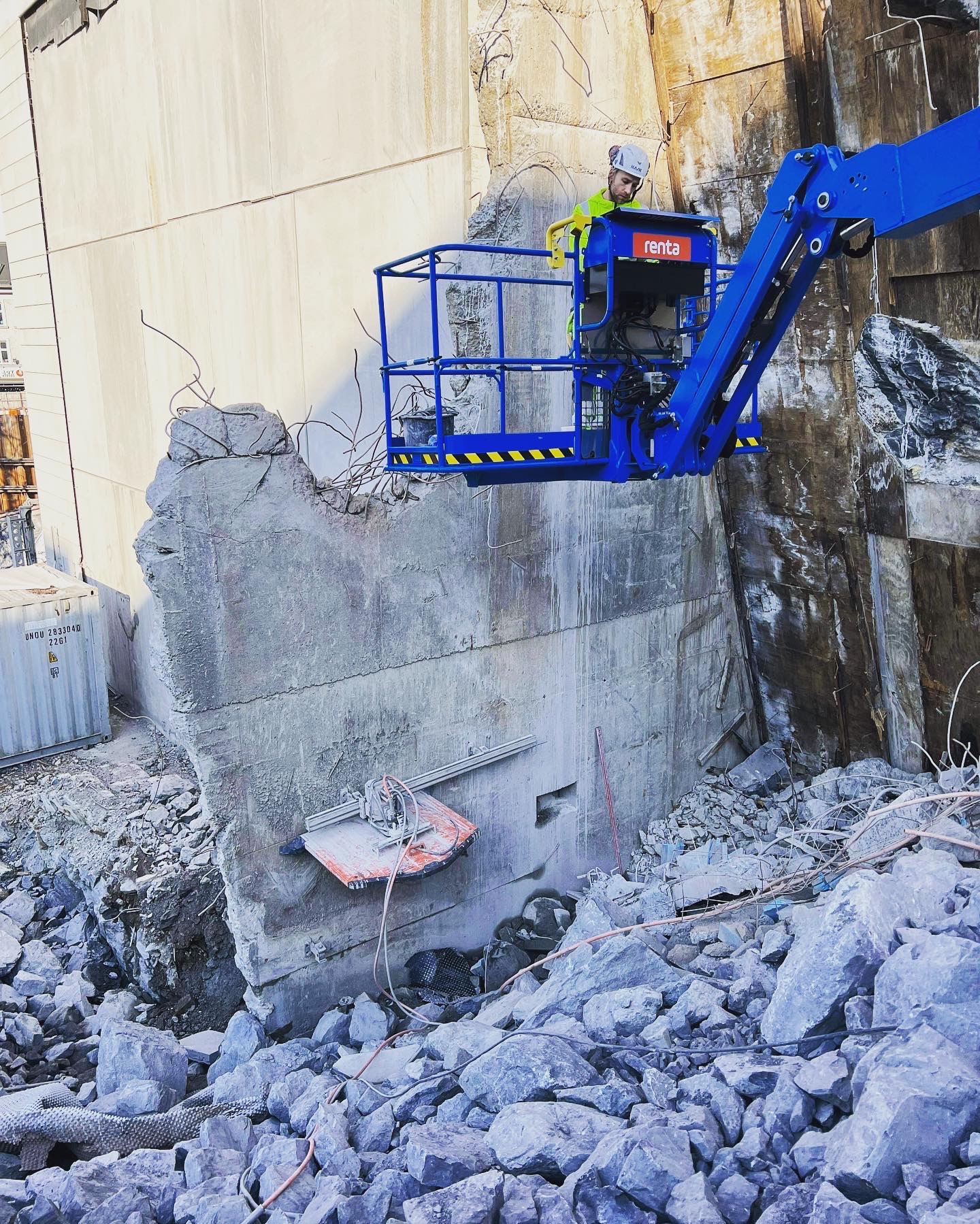 Pracownik na podnośniku Renta wyburza ścianę z betonu zbrojonego za pomocą piły diamentowej. Wokół gruz i fragmenty zbrojenia. Widok z dołu.