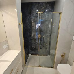 Geblon - Nowoczesna łazienka z kabiną prysznicową typu walk-in, wykończoną złotymi detalami. Marmurowe płytki w odcieniach szarości i bieli. Biała szafka z umywalką i złotymi uchwytami.