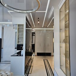 Geblon - Eleganckie wnętrze: błyszczący sufit z oświetleniem LED, okrągły żyrandol, białe meble z złotymi akcentami i marmurowa podłoga z czarnymi pasami.