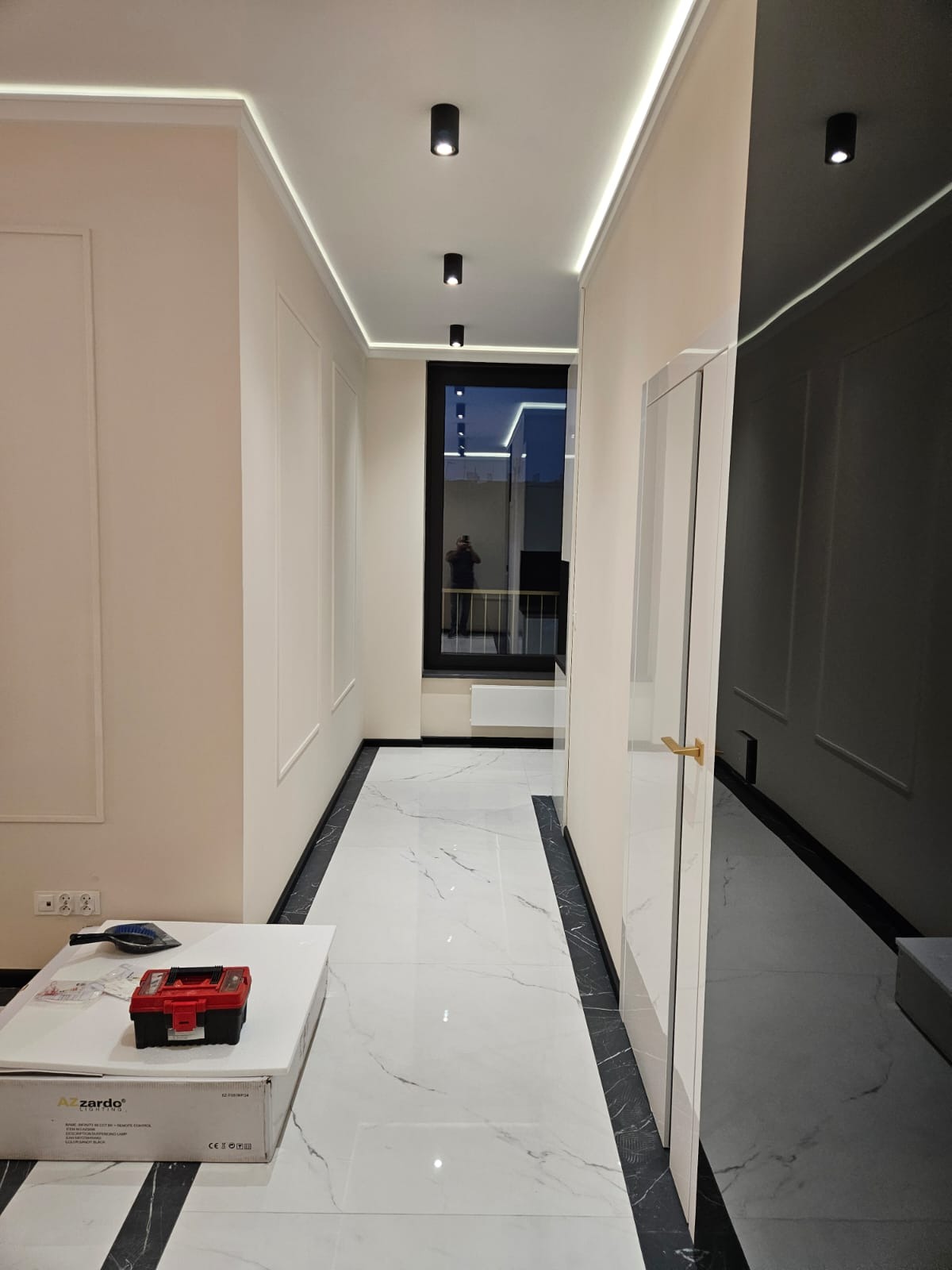 Wykończony korytarz z białymi ścianami, czarnymi lampami sufitowymi i podłogą z białego marmuru z czarnymi obramowaniami. Widoczne narzędzia na stole.