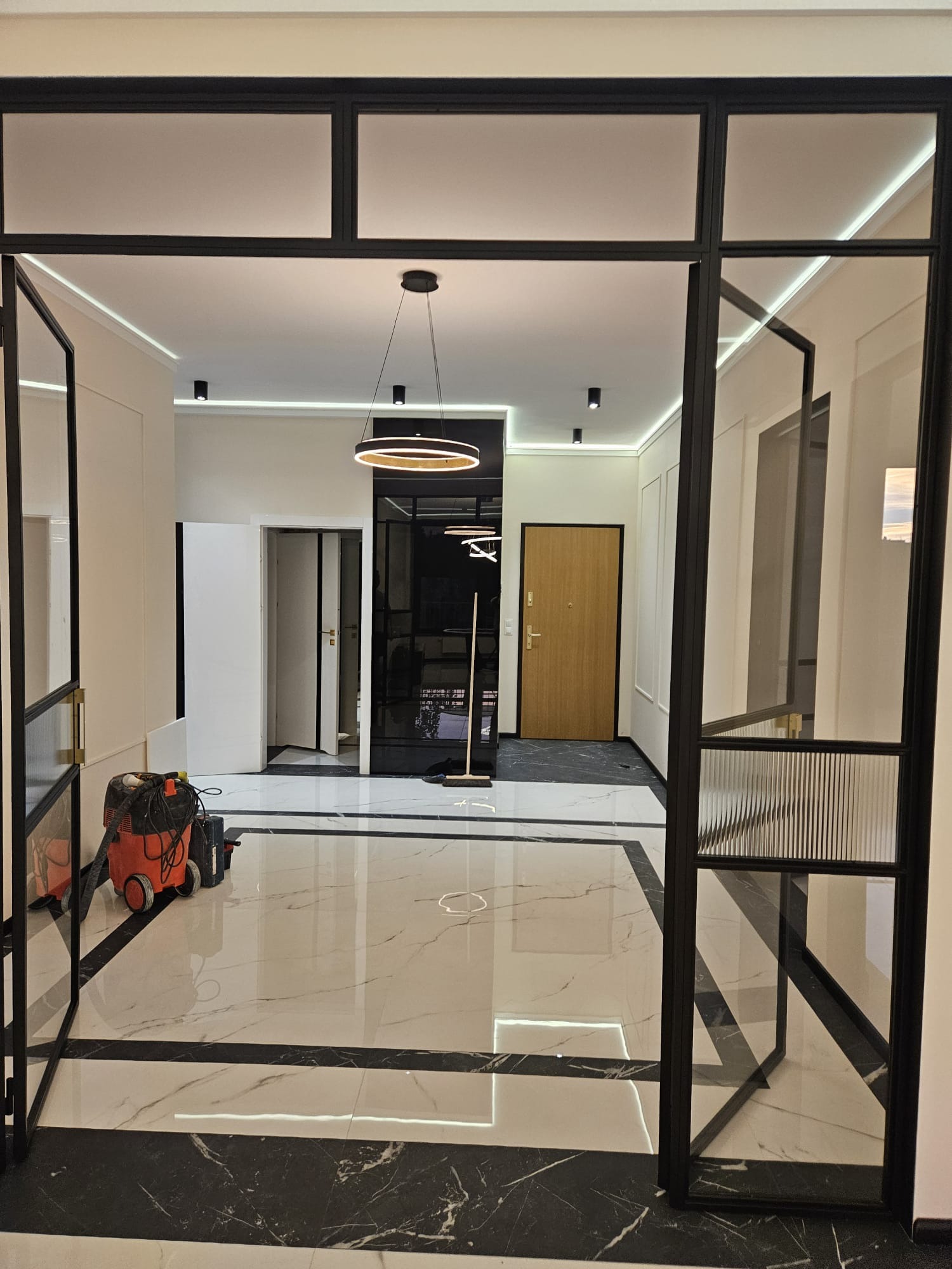 Nowoczesny hol z otwartymi, czarnymi, przeszklonymi drzwiami. Widoczna marmurowa podłoga w biało-czarne wzory i wisząca lampa. Odkurzacz na podłodze.