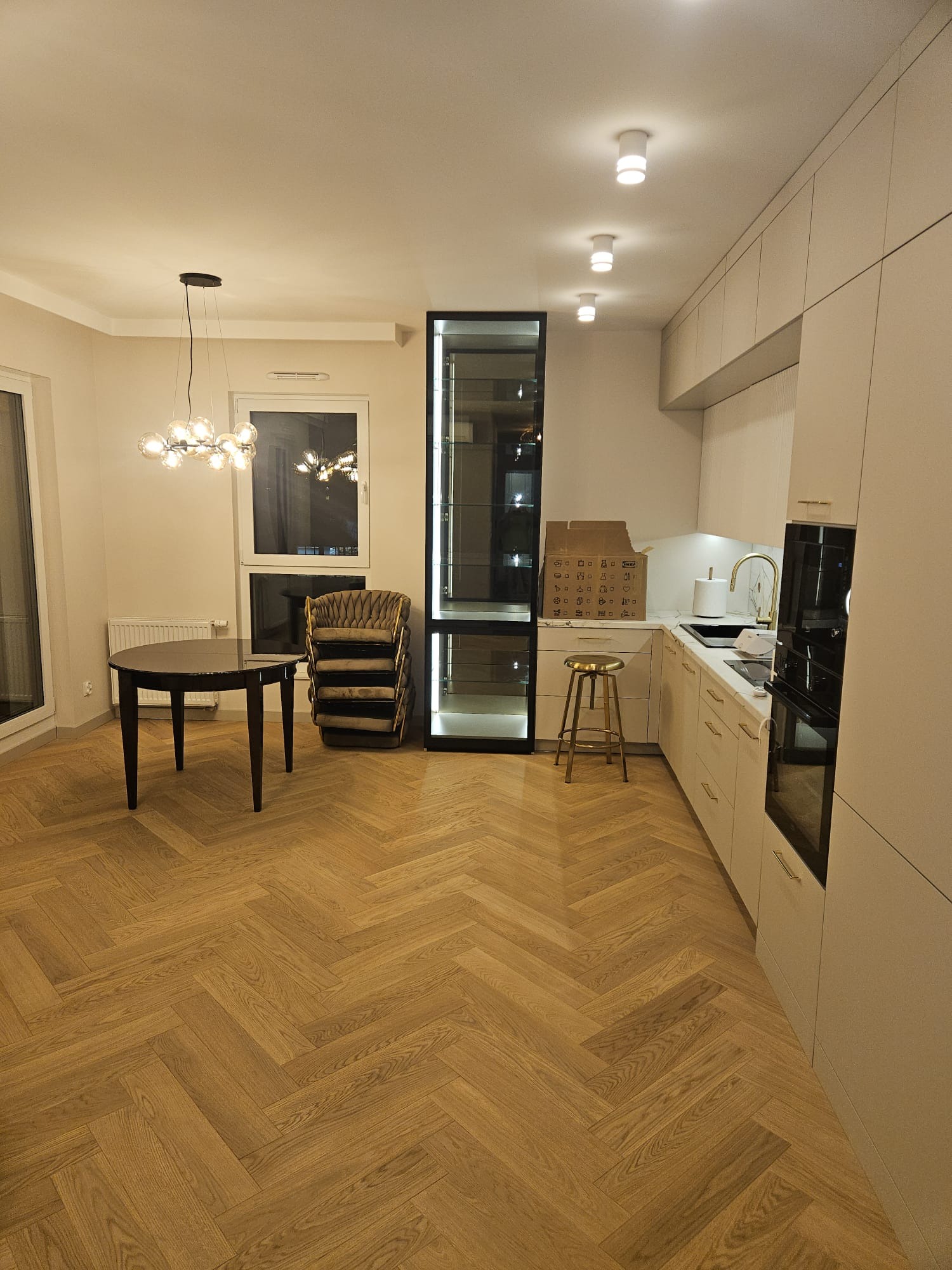 Jasna kuchnia z parkietem w jodełkę, oświetleniem punktowym i wiszącą lampą. Widoczne krzesła w rogu i wysoka witryna. Minimalistyczny design.