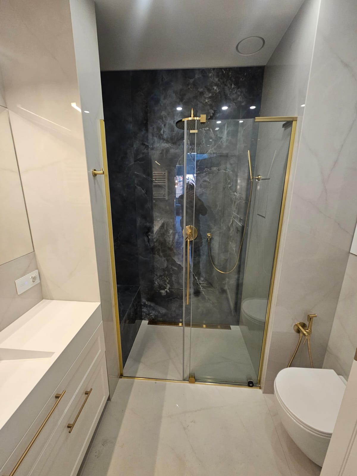 Nowoczesna łazienka z kabiną prysznicową typu walk-in, wykończoną złotymi detalami. Marmurowe płytki w odcieniach szarości i bieli. Biała szafka z umywalką i złotymi uchwytami.