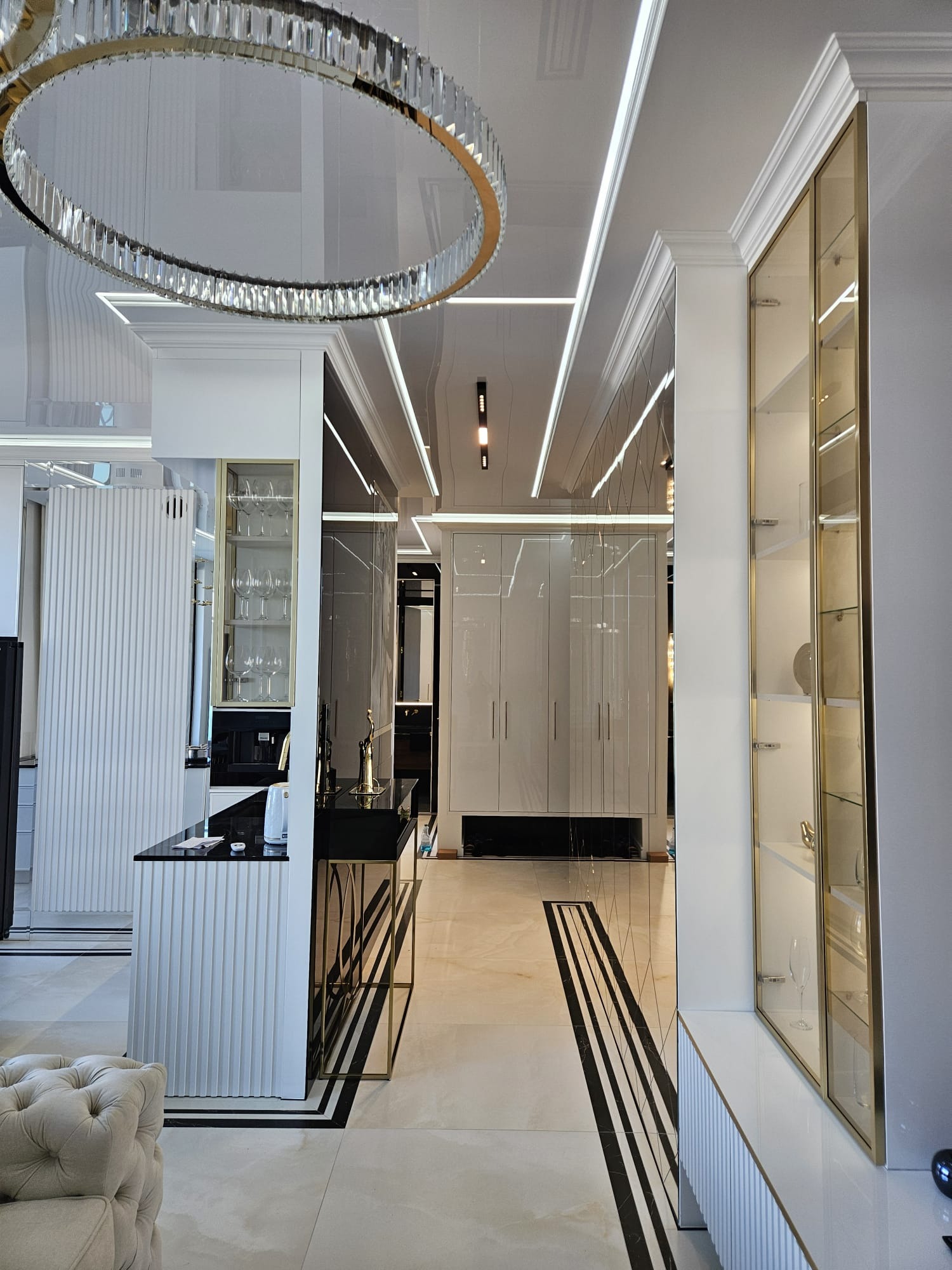 Eleganckie wnętrze: błyszczący sufit z oświetleniem LED, okrągły żyrandol, białe meble z złotymi akcentami i marmurowa podłoga z czarnymi pasami.