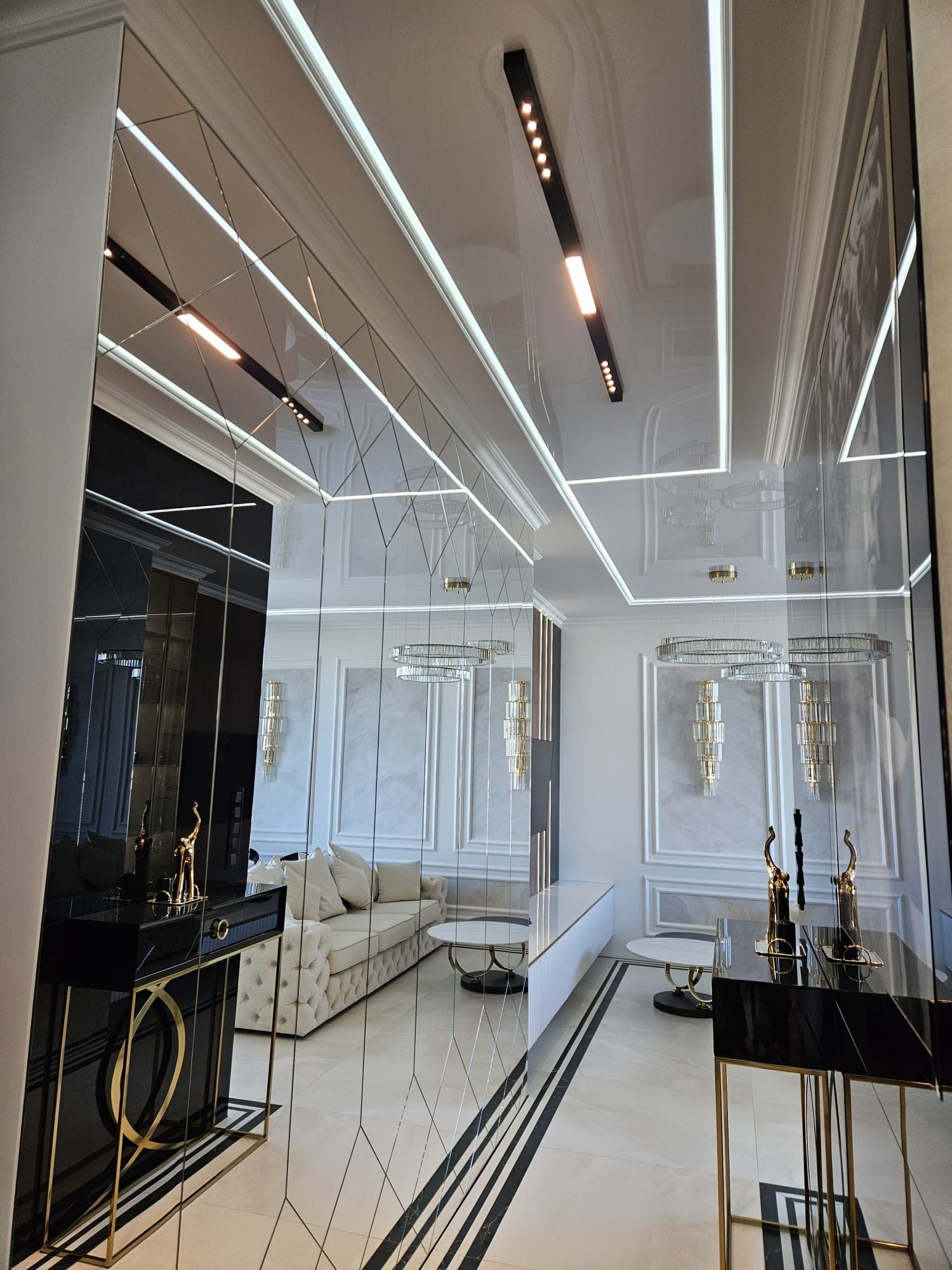 Eleganckie wnętrze salonu z lustrzanymi ścianami, oświetleniem LED i dekoracyjnymi lampami wiszącymi. Widoczny fragment sofy i stolików kawowych w jasnej kolorystyce.