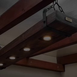 Mariusz isiu - Drewniana lampa wisząca z czterema punktami świetlnymi, zawieszona na łańcuchach, w rustykalnym stylu, z widoczną strukturą drewna i metalowymi elementami.