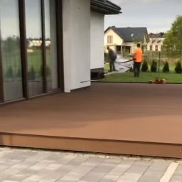 Mariusz isiu - Nowoczesny taras z deski kompozytowej w Żarówce. Widoczne prace wykończeniowe i fragment domu z dużymi oknami. Minimalistyczny design i estetyczne wykończenie.